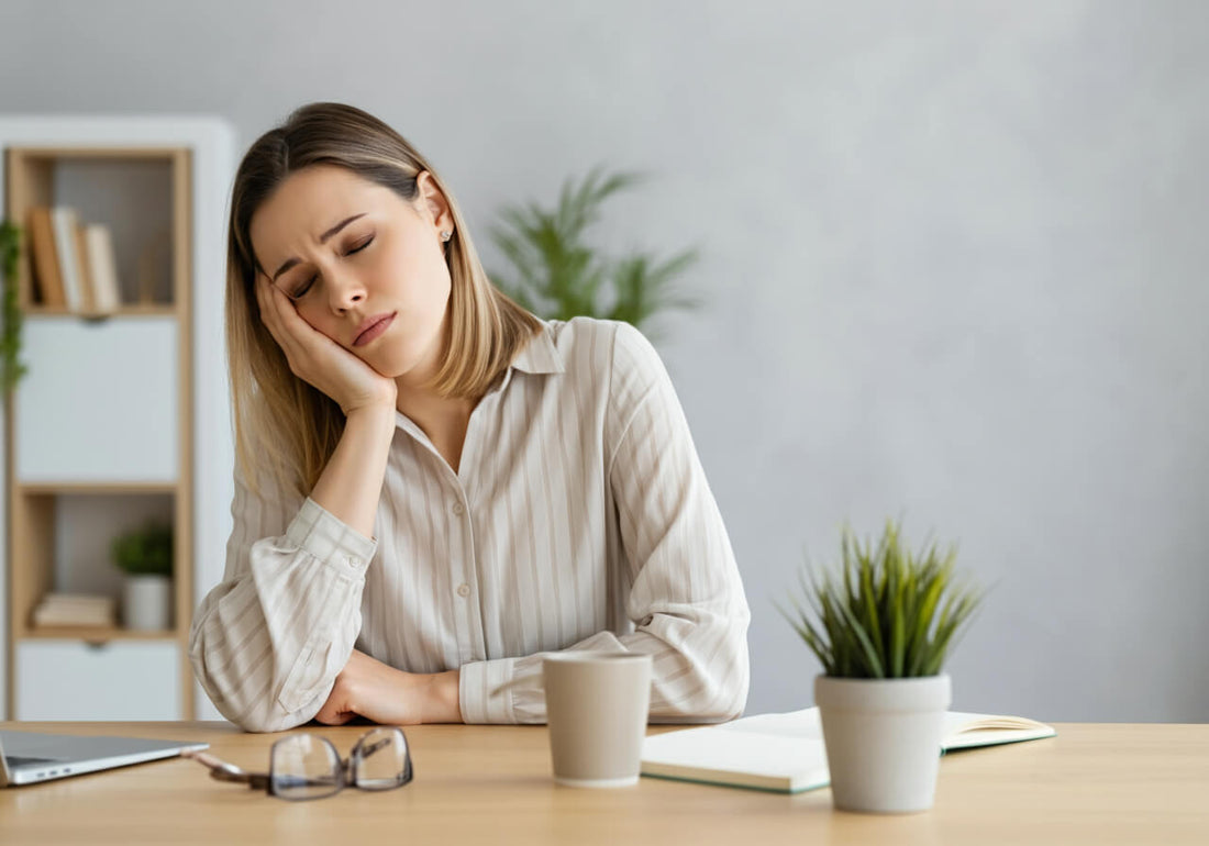 A woman wearing a grayish button-up shirt with long sleeves and collar. She is in an office with gray pain. She has her right hand on her right cheek. She is sleepy, her eyes are closed.