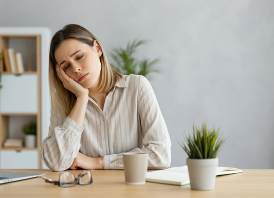 A woman wearing a grayish button-up shirt with long sleeves and collar. She is in an office with gray pain. She has her right hand on her right cheek. She is sleepy, her eyes are closed. 