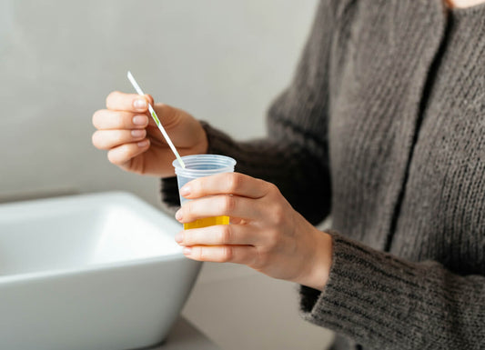 A woman in gray sweater dipping a urine test strip on a cup of water. She is inside a bathroom, with the sink nearby