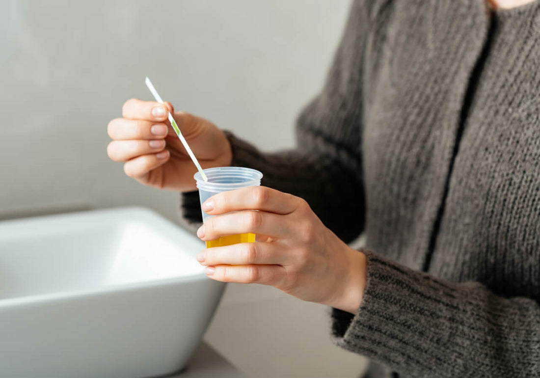 A woman in gray sweater dipping a urine test strip on a cup of water. She is inside a bathroom, with the sink nearby